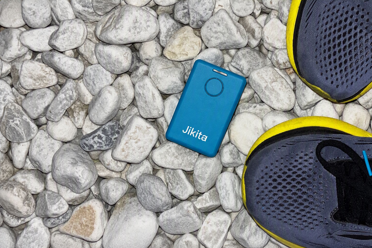 A blue Jikita beacon lying on white pebbles beside a pair of trail-running shoes